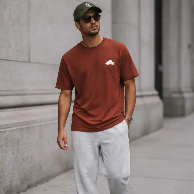 Man wearing a brown t-shirt, gray pants, and a cap walking on a city street.